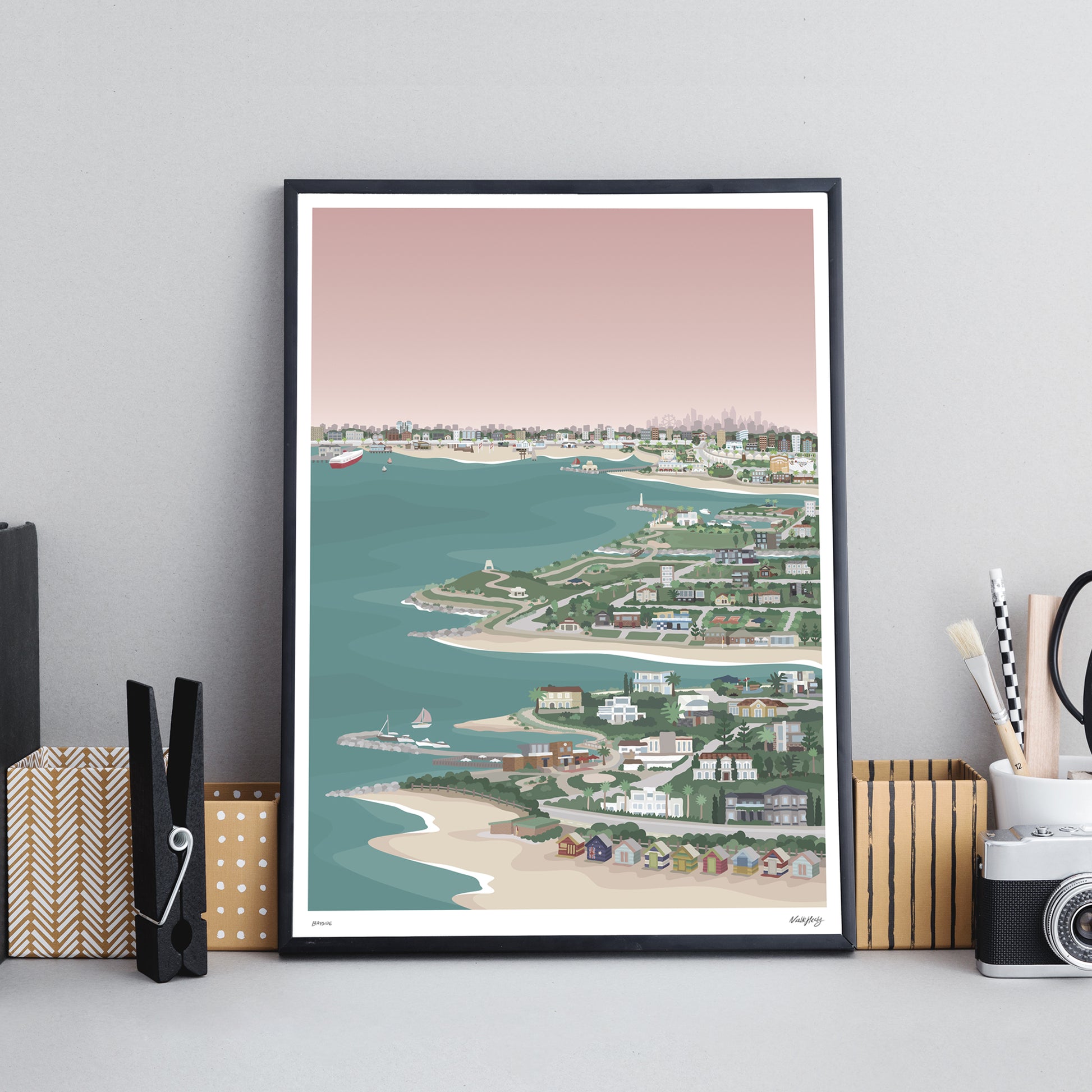 black framed colourful art print of bayside Melbourne from Princes pier to Brighton bathing boxes displayed on an office desk